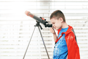 Child wearing a red cape and looking through a telescope on a tripod out of a large window.
