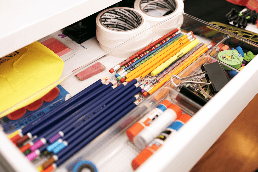 Close up of open desk drawer containing materials for a child to use in creative activities. Drawer contents include masking tape, colored pencils, scissors, glue sticks, and paper clips. 