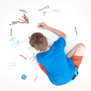 Overhead view of child sitting cross legged bent over and coloring with crayons on a large sheet of white paper.