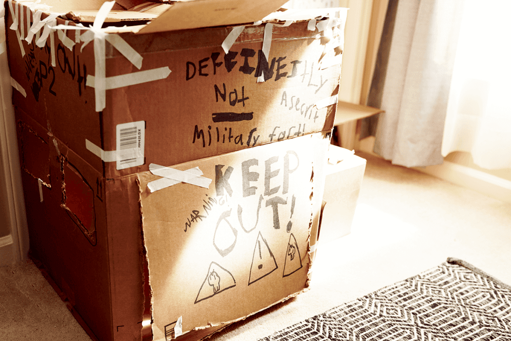 Cardboard fort with child's drawings on it such as "Keep out" and "Secret fort". Cardboard box is held together with white masking tape.