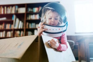 little girl playing pretend in an astronaut helmet while coloring with crayons on a cardboard box