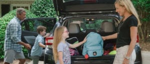 mom helping daughter put her backpack in the trunk of a car. A brick red relay with wearable loop case is clipped to the backpack.