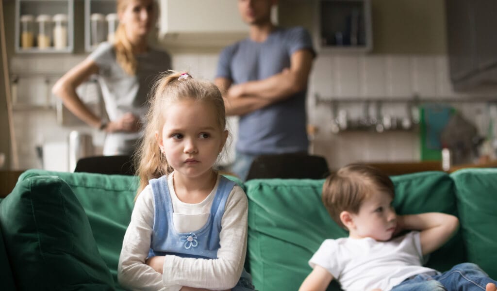girl sits with her brother on the couch. She ignores her parents, who observe her behavior.